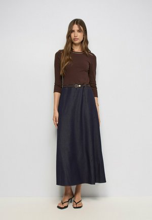 Mujer con camiseta marrón de manga larga, falda midi azul oscuro de línea A con cinturón y sandalias negras de tacón, de pie frente a un fondo blanco.