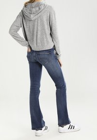 Sudadera corta gris con capucha ajustable, combinada con unos vaqueros acampanados azules y zapatillas blancas con rayas negras. Vista posterior mostrada.