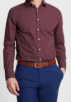 Mann trägt ein bordeauxrotes, gemustertes Hemd, in blaue Hosen gesteckt, mit einem braunen Ledergürtel, eine Hand in der Tasche.