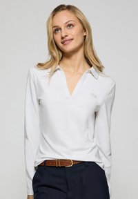 Vit långärmad polo med v-ringning, med en diskret logotyp på vänster bröst. Modellen bär marinblå byxor och ett brunt bälte.