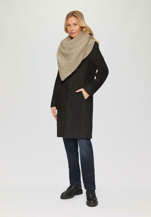 Manteau en laine noire, mi-long, avec deux poches latérales. Écharpe en tricot beige à motifs drapée autour du cou. Jean noir et bottes en cuir.