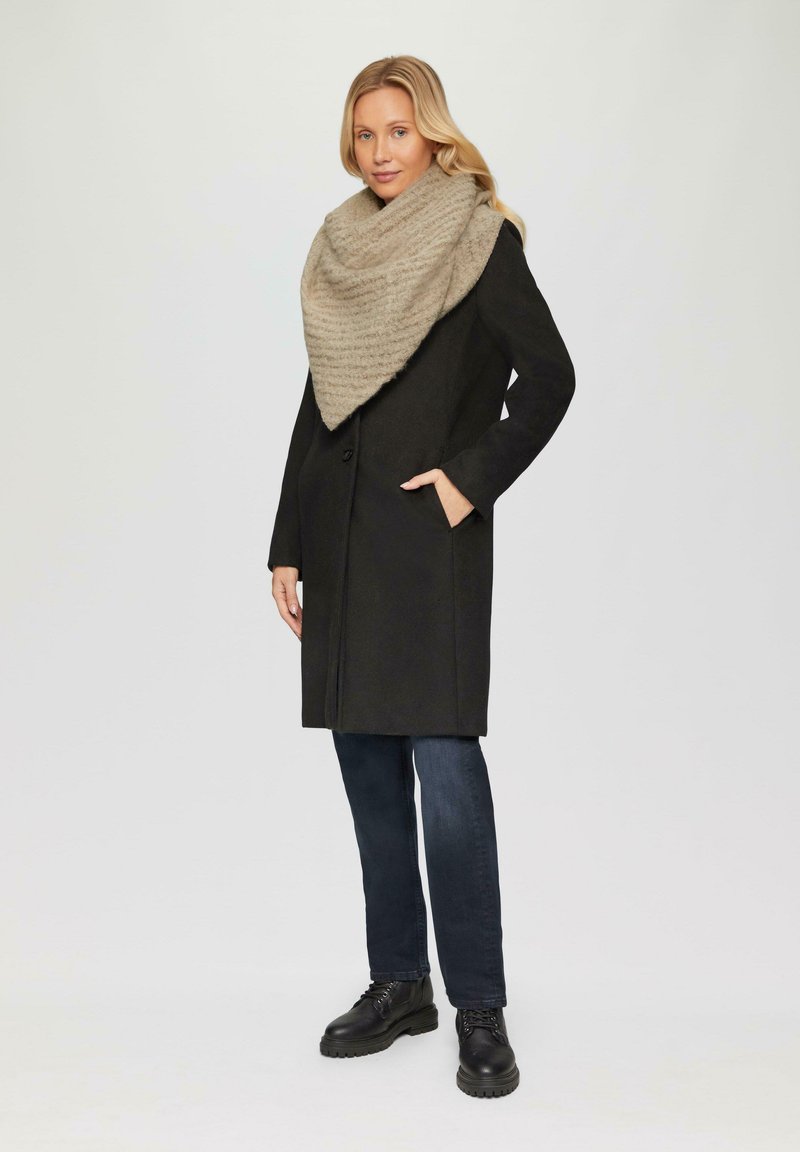 Manteau en laine noire, mi-long, avec deux poches latérales. Écharpe en tricot beige à motifs drapée autour du cou. Jean noir et bottes en cuir.