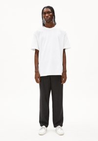 Weißes Baumwoll-T-Shirt, lockere Passform mit kurzen Ärmeln, kombiniert mit dunklen, locker sitzenden Hosen und weißen Sneakers, vor einem schlichten Hintergrund stehend.