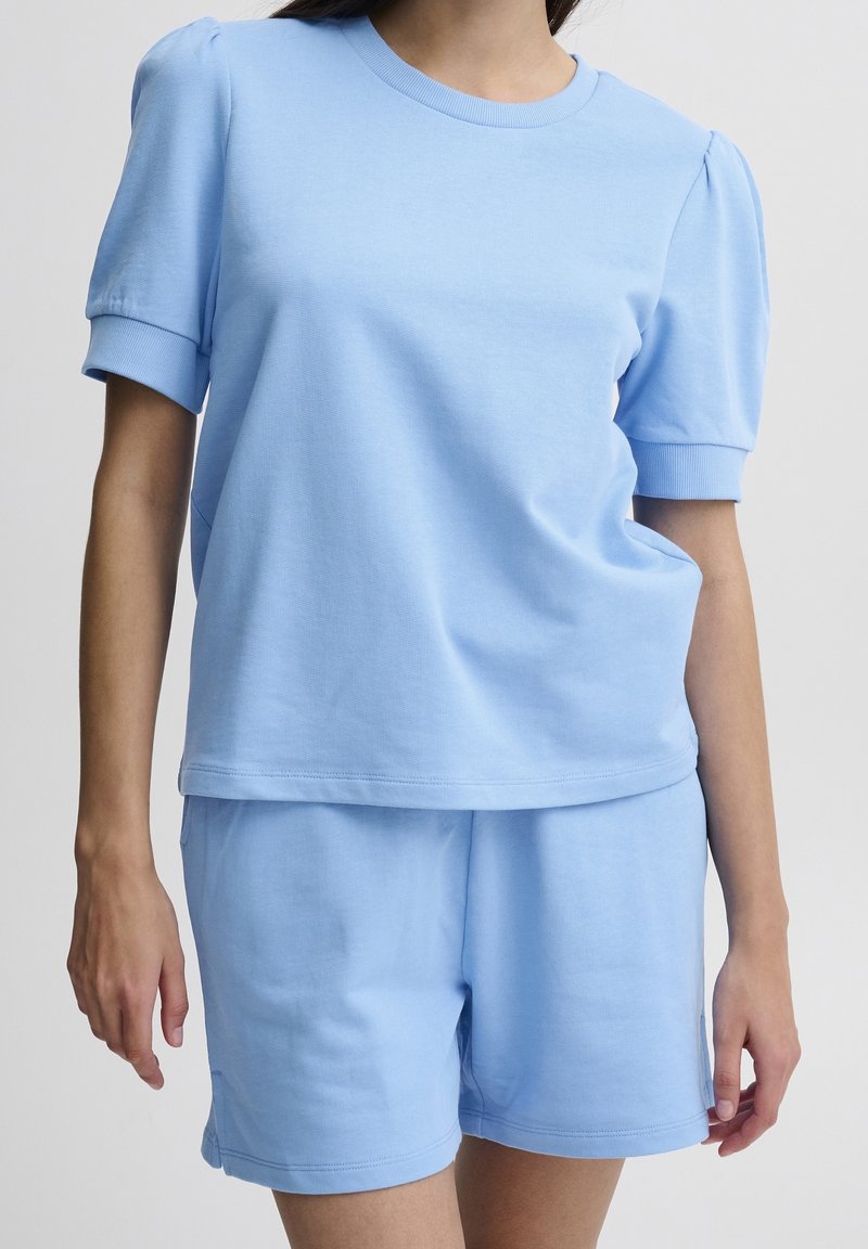 Kvinna som bär en ljusblå kortärmad skjorta med puffiga axlar och matchande lösa shorts mot en enfärgad bakgrund.