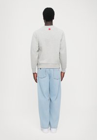Lys grå bomuldssweatshirt med ribbede manchetter og kant, prydet med et lille rødt logo på ryggen. Parret med lyseblå baggy jeans.