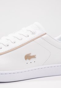 Baskets blanches avec logo crocodile beige, détail de bordure beige, lacets blancs et semelle blanche, montrant le côté et la zone supérieure des lacets.