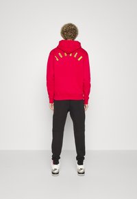 Rote Kapuzenjacke mit "JORDAN" in Gelb auf dem Rücken, kombiniert mit schwarzen Hosen und weißen Sneakers in niedrigem Design.