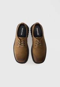 Ruskohvaiset mokkasiinit, joissa on pyöreä kärki, musta kumipohja ja litteät nauhat. Logo näkyy sisäpohjassa. Sileä pinta, jossa on liukuväri-efekti.