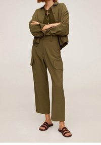 Tuta cargo verde oliva con giacca colletto, caratterizzata da grandi tasche, chiusura con bottoni e sandali scivolati marroni. Texture del tessuto morbida.