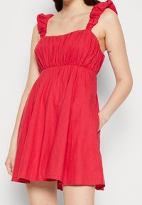 Robe rouge sans manches avec des bretelles froncées, un corsage froncé et une jupe fluide. Fabriquée en tissu léger, elle dispose de poches latérales et d'une texture douce.