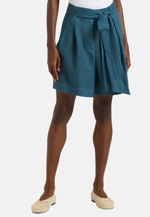 Donna che indossa pantaloncini a vita alta color verde acqua con pieghe e cintura di tessuto annodata, abbinati a ballerine beige intrecciate, in piedi davanti a uno sfondo semplice.