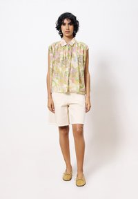 Mouwloos, pastelbloemig shirt met een kraag, gecombineerd met crèmekleurige shorts. Het shirt heeft een losse pasvorm en een gestructureerde stof. Bruine platte schoenen.