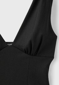 Gros plan d'un vêtement sans manches noir texturé avec des larges bretelles et un détail de couture froncée sous l'épaule.