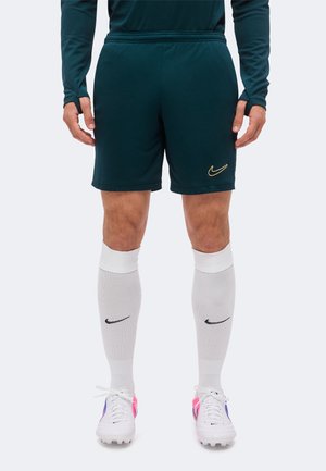 Hombre que lleva pantalones cortos deportivos verde oscuro, calcetines blancos hasta la rodilla con logo negro y botas blancas con detalles en rosa y púrpura.