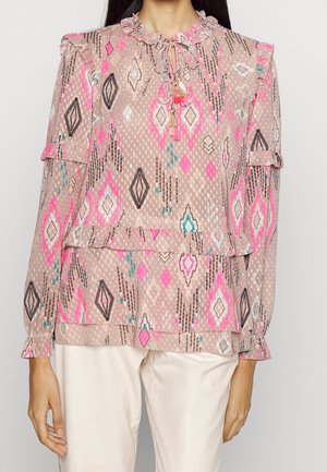 Femme portant un chemisier rose à manches longues avec des motifs géométriques, un col volanté et un détail noué, associé à un pantalon de couleur claire.
