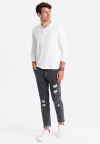 Hombre con polo blanco de manga larga, jeans oscuros desgastados enrollados en los tobillos, zapatillas blancas y pulseras negras de cuentas, mirando hacia su izquierda.