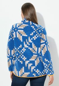 Kurtka polarowa z niebieskim, beżowym i białym geometrycznym wzorem, z wysokim kołnierzem i pomarańczowym obszyciem na krawędzi. Miękka w dotyku.