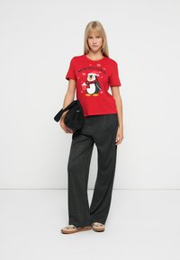 Rød kortærmet t-shirt med et pingvinmotiv, parret med grå bukser med bred ben og beige slip-on sko. Sort taske i hånden.