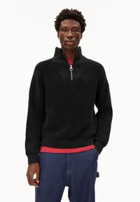 Mann, der einen schwarzen Strickpullover mit Reißverschluss über einem roten Hemd und blauen Hosen trägt, steht mit einer Hand in der Tasche vor einem weißen Hintergrund.