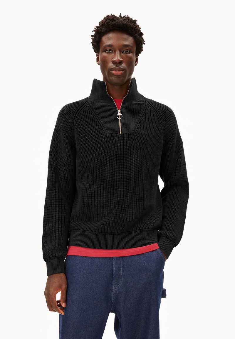 Mann, der einen schwarzen Strickpullover mit Reißverschluss über einem roten Hemd und blauen Hosen trägt, steht mit einer Hand in der Tasche vor einem weißen Hintergrund.