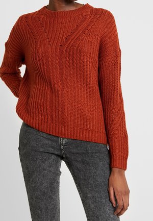 Pull en maille côtelée couleur rouille à manches longues porté avec un jean noir délavé sur une personne debout devant un fond uni.