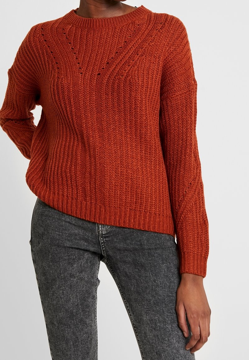 Pull en maille côtelée couleur rouille à manches longues porté avec un jean noir délavé sur une personne debout devant un fond uni.