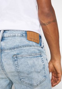 Jeans de mezclilla azul claro con un acabado texturizado, que presentan bolsillos traseros y un parche de cuero marrón con la etiqueta "American Eagle" en la cintura.