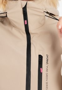 Veste beige avec fermetures éclair étanches noires, tirettes de fermeture éclair roses, et texte "protest / PVRE GREEN SERIES", mains ajustant les cordons de la capuche.