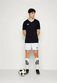 Melnā sporta krekls ar baltajiem akcentiem, apvienots ar baltām šortiem. Modelis stāv ar melnbaltu futbola bumbu pie kājām, ģērbies melnās futbolu kurpēs.