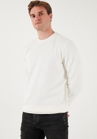 T-shirt bianco a maniche lunghe con scollatura rotonda, texture a coste e orlo dritto. Indossato con jeans scuri.