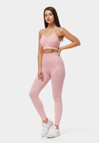 Soutien-gorge de sport rose clair avec un devant froncé et un legging taille haute assorti, présentant une texture lisse et un branding subtil. Porté avec des baskets blanches.