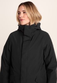 Woman with light blonde bob hairstyle wearing a black hooded winter jacket, looking to the left against a plain background.