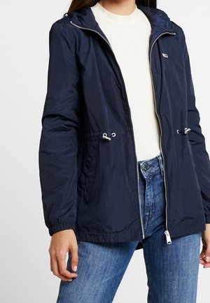Kvinde iført en mørkeblå hættejakke med lynlås og snører, en hvid ribstrikket sweater og blå jeans.