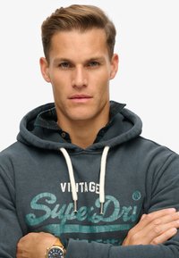 Donkerblauwe hoodie met witte trekkoorden en opdruk. Gelaagd met een groene geruite kraag. Gladde stofstructuur, casual ontwerp.