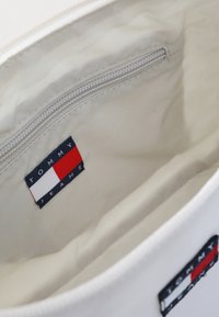 Interior de una bolsa de tela blanca que cuenta con un bolsillo con cremallera y una etiqueta azul marino con un logotipo en rojo, blanco y azul que dice "TOMMY JEANS." Textura suave.