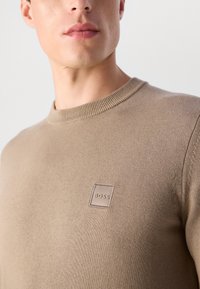 Közelkép egy személyről, aki bézs kötött pulóvert visel, a mellén egy kis "BOSS" logóval, egyszínű háttérrel.