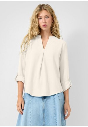 Femme blonde aux cheveux ondulés portant un chemisier crème à col en V avec manches retroussées et une jupe en jean bleu clair sur un fond uni.