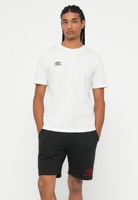 Mladenič z pletenicami, oblečen v belo majico Umbro in črne kratke hlače Umbro, stoji z rokama v žepih pred enobarvnim ozadjem.