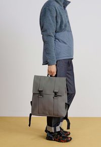 Sac à dos gris au design structuré, avec des doubles bretelles à l'avant et une finition mate. Porté par une personne en veste polaire bleue et pantalon sombre.