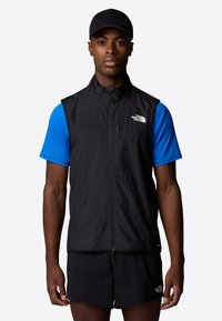 Black lightweight vest with a high collar and front zipper, worn over a short-sleeve blue shirt, paired with black shorts. Branding visible.