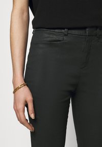 Black slim-fit pants with a smooth texture and a slight sheen, paired with a gold chain bracelet on a light skin hand.