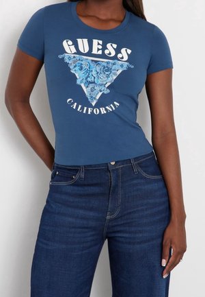 Kvinde iført en tætsiddende blå GUESS California T-shirt med pailletblomstret trekantdesign, kombineret med højtaljede blå jeans.