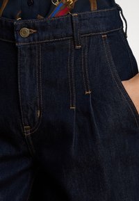 Mørkeblå denimjeans med strukturert midje, kontrastsøm og frontfolder. Har en metallknapp og flere lommer.