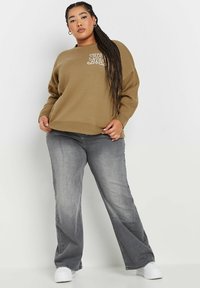 Khaki Pullover mit lässigem Schnitt, gerippten Bündchen und weißen Grafiken; kombiniert mit ausgebleichten grauen Jeans und weißen Sneakers.