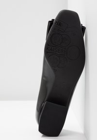 Semelle de chaussure noire polie avec des motifs de pièces en relief et le mot Kaiser, vue en angle sur fond blanc.