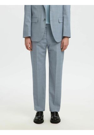Personne portant un pantalon de costume à carreaux bleu clair, un blazer assorti, un haut bleu clair et des mocassins en cuir noirs sur fond blanc.