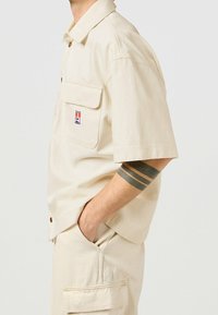 Camisa de manga corta beige con cierre de botones, dos bolsillos en el pecho y un parche con el logo. Fabricada con un tejido suave y ligeramente texturizado.