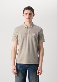 Beige polo skjorte med kragehals, korte ermer og en subtil logo patch på venstre bryst. Kombinert med blå denim jeans.