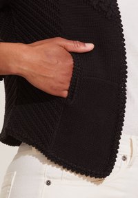 Černá pletená bunda s texturovanou tkaninou, otevřeným předním dílem a boční kapsou. Ozdobená vlnitým okrajem podél lemu.