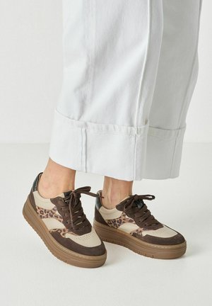 Feet wearing white pants and brown sneakers with leopard print and thick rubber soles on a white floor.